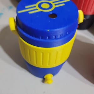 May include: A blue and yellow plastic cooler-shaped container featuring a yellow Fallout Vault-Tec logo on the lid. The cylindrical object has a yellow band around the center, a small yellow spigot at the base, and two yellow side handles.
