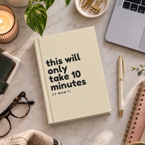 May include: A cream-coloured hardback notebook with the black text "this will only take 10 minutes (IT WON'T)" printed on the front cover. The notebook is surrounded by office supplies including a pen, glasses, and a laptop on a marble surface.