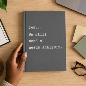 Op de afbeelding: Een donkergrijs notitieboek met harde kaft en witte tekst op de voorkant die luidt: Yes... We still need a needs analysis. Het notitieboek is rechthoekig met een effen matte afwerking en een verticale rug aan de linkerkant van de kaft.