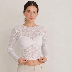 May include: A long-sleeved white lace top featuring a sheer floral pattern with a built-in opaque sweetheart-style bodice lining. The garment is paired with solid beige tailored shorts that have a flat front, a waistband, and side pockets.