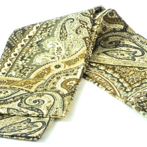 May include: A folded scarf with a paisley pattern in shades of brown, olive green, and cream. The scarf has a detailed design with curved, teardrop-shaped motifs and floral elements. The fabric appears to be soft and flowing.