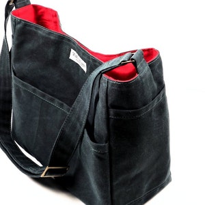 Pode incluir: Uma bolsa tote de lona cinza escura com forro vermelho e vários bolsos. A bolsa tem uma alça longa ajustável e um pequeno rótulo que diz "Feito no Canadá".
