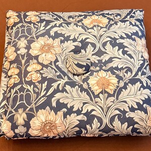 Peut inclure: Un coussin de siège carré présentant un fond bleu foncé avec un motif répétitif de fleurs beige clair et un feuillage complexe. Le coussin comprend un bouton unique recouvert de tissu centré sur la surface supérieure, offrant un aspect capitonné.