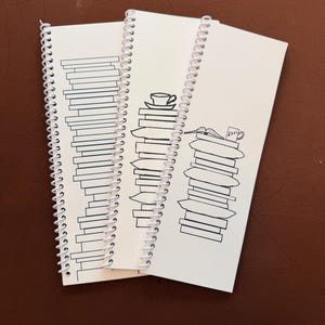 May include: Three white spiral-bound notebooks featuring black line art illustrations. The first shows a tall stack of books, the second a stack of books topped with a teacup, and the third a stack of books with an open book and a mug labeled ZZZ.