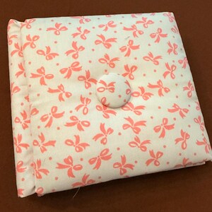 Peut inclure: Un coussin carré capitonné présentant une base en tissu crème avec un motif répétitif de petits nœuds et de points rose corail. Le centre du coussin est agrémenté d'un bouton unique recouvert du même tissu à motif de nœuds.