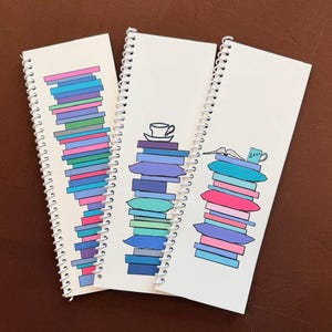 May include: Three white spiral-bound notebooks featuring colorful illustrations. The first shows a tall stack of books in shades of blue, pink, and purple. The second displays a stack of pillows topped with a teacup. The third shows pillows with a book and mug.