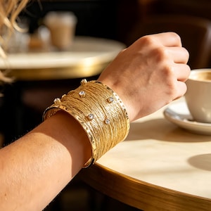 Può includere: Un ampio bracciale a fascia color oro caratterizzato da una superficie testurizzata simile a fili intrecciati e diverse piccole pietre chiare a taglio rotondo incastonate sul davanti. Il bracciale presenta bordi lisci e una struttura rigida.