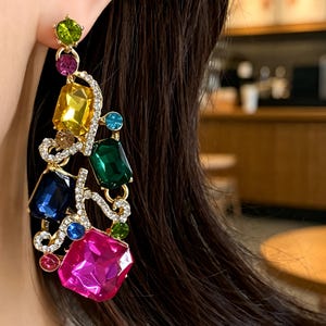 Puede incluir: Un pendiente largo colgante con una estructura de metal dorado adornada con cristales facetados multicolores en tonos rosa, amarillo, verde, azul y verde oscuro, acentuado por pequeños brillantes transparentes en un diseño de filigrana.