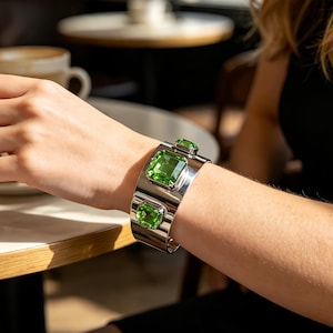 Puede incluir: Una pulsera de brazalete ancha en tono plateado con tres grandes piedras preciosas rectangulares de color verde facetado engastadas en metal pulido. La pulsera tiene una superficie lisa y reflectante y se lleva en la muñeca.