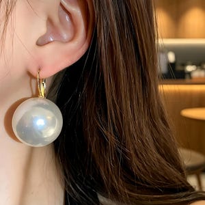 May include: A large, spherical white pearl earring featuring a smooth, lustrous surface and a reflective finish. The pearl is suspended from a gold-toned metal lever-back clasp that secures the earring to the earlobe with a simple, functional design.