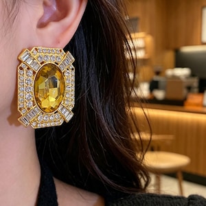 May include: A large rectangular gold-tone earring featuring a central oval-shaped amber-colored gemstone. The frame is decorated with a border of small round clear crystals and baguette-cut clear crystals arranged in a geometric pattern.
