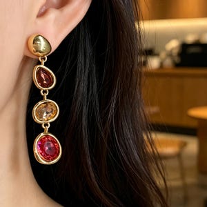 Puede incluir: Un pendiente colgante de tono dorado con un perno redondeado y liso en la parte superior, seguido de tres dijes colgantes de forma irregular. Los dijes contienen piedras facetadas y translúcidas en tonos rojo oscuro, ámbar y rosa brillante, engastadas en oro.