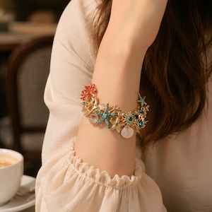 May include: A gold-tone charm bracelet featuring various sea-themed elements including starfish, seashells, and coral-like structures. The charms are embellished with small blue, clear, and red crystals, creating a detailed ocean-inspired accessory.