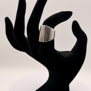 May include: A wide, smooth, silver-colored metal ring with a curved, shield-like front surface. The band features a polished, reflective finish that tapers slightly toward the back, creating a solid and continuous metallic loop design.