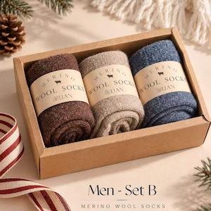 Puede incluir: Un conjunto de tres pares de calcetines de lana merino en marrón, beige y azul, empaquetados en una caja de cartón rectangular. Cada par está envuelto en una banda de papel con la etiqueta MERINO WOOL SOCKS LIAN y un icono de oveja.