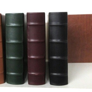 Könnte beinhalten: Eine Sammlung von fünf ledergebundenen Büchern in verschiedenen Farben: Gelb, Orange, Grün, Burgunderrot und Schwarz. Eine passende braune Box befindet sich neben den Büchern. Die Buchrücken haben horizontale Bänder.