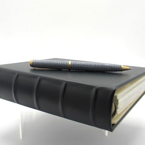 May include: A black leather journal with a silver and gold pen on top. The pen has a textured grip and gold detailing. The journal's pages are visible, and the spine has a classic, raised-band design.