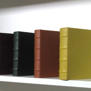 May include: Five leather-bound books on a white shelf. The books are in different colors: black, dark brown, green, brown, and yellow.