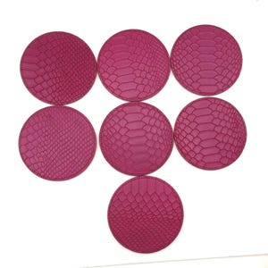 May include: Seven magenta-coloured, round coasters with a textured, snakeskin-like pattern. The coasters are arranged in a circular pattern against a white background. The coasters are made of a durable material.