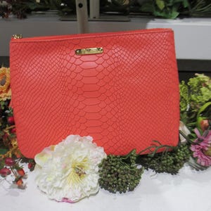 May include: A rectangular coral-colored clutch bag featuring a textured snakeskin-like pattern. The bag has a gold-tone zipper closure along the top edge and a small, rectangular gold-tone metal plaque centered near the top with engraved text.
