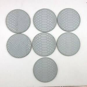 May include: Seven round, gray coasters with a snakeskin pattern. The coasters are arranged in a circular pattern. Each coaster has a stitched edge. The coasters are made of a durable material.