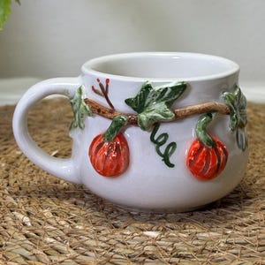 Könnte beinhalten: Ein weißer Keramikbecher mit einem erhabenen, dreidimensionalen Design aus orangefarbenen Kürbissen, grünen Blättern und braunen Ranken, die sich um die Außenseite winden. Der Becher hat eine glatte, glänzende Oberfläche und einen geschwungenen Henkel.