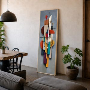 May include: A tall, rectangular framed wall art piece featuring a vertical composition of layered geometric shapes in shades of red, orange, yellow, teal, blue, and cream against a textured gray background, displayed leaning against a wall.