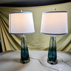 May include: A pair of table lamps featuring tapered, ribbed glass bases that transition from a deep teal at the bottom to a lighter clear hue at the top. Each lamp is topped with a white drum shade and sits on a circular brushed metal base.
