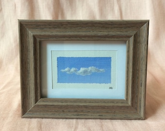 Op de afbeelding: Een ingelijst schilderij van een enkele witte wolk tegen een blauwe lucht. De lijst is van lichtbruin hout.