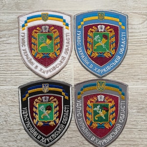 May include: Four shield-shaped embroidered patches featuring a central emblem with a cornucopia and caduceus on a green background, surrounded by a maroon field and a blue and yellow border. Each patch includes Ukrainian text and a small trident symbol.