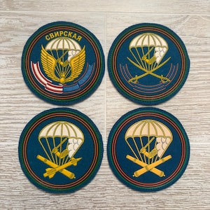 May include: Four circular embroidered patches on a light wood surface. Each features a white parachute and yellow emblem. The top left patch includes the Cyrillic text "СВИРСКАЯ". The others display crossed swords, missiles, or cannons beneath the parachute.