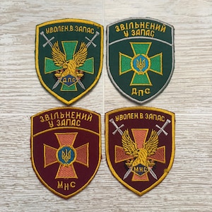 Pode incluir: Quatro emblemas bordados em forma de escudo com designs militares. Dois são verdes com bordas amarelas e dois são bordô com bordas amarelas. Exibem texto cirílico, espadas cruzadas, águias e o símbolo do tridente ucraniano.