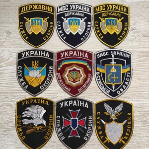 May include: A collection of nine embroidered patches featuring various Ukrainian state security and service emblems. The shield-shaped patches display intricate designs including the Ukrainian trident, swords, eagles, and Cyrillic text in black, gold, blue, and red.