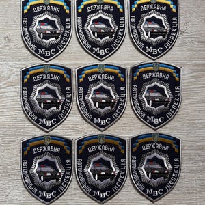 May include: Nine shield-shaped embroidered patches featuring a dark blue background, a central police car graphic, and the Cyrillic text "ДЕРЖАВНА АВТОМОБІЛЬНА ІНСПЕКЦІЯ МВС". Each patch includes a blue and yellow ribbon design at the top and a silver border.