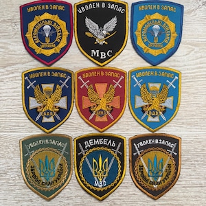 May include: A collection of nine shield-shaped embroidered patches featuring various military-themed designs, including eagles, parachutes, swords, and the Ukrainian trident symbol, with Cyrillic text such as Uvolen v zapas and MVS.
