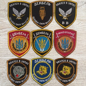 May include: A collection of nine shield-shaped embroidered patches featuring various designs including eagles, owls, tigers, and the Ukrainian trident symbol. The patches display Cyrillic text and are bordered in yellow, red, or blue thread.