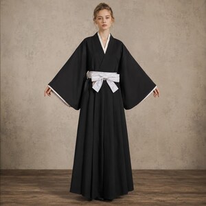 May include: A black floor-length robe with wide sleeves and a white collar trim. The garment features a white sash tied in a bow at the waist, creating a structured silhouette. The design includes a long, pleated skirt section that falls to the floor.