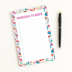 May include: A white notepad with a colorful floral border and the text "MARISSA FLORES" in pink. The notepad has a lined interior with check boxes. A black pen with gold accents rests beside the notepad.