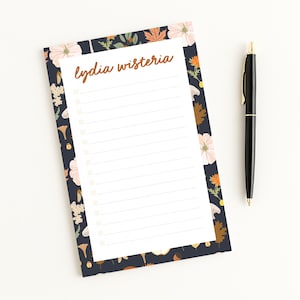 May include: A notepad with a floral border and the text "lydia wisteria" in a handwritten style. The notepad has a dark blue background with white flowers, orange accents, and brown leaves. A black pen with gold accents rests beside the notepad.