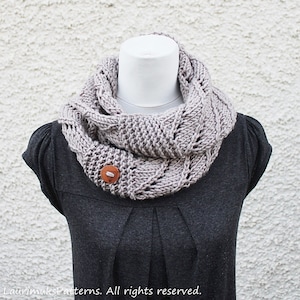 Op de afbeelding: Een grijze gebreide oneindige sjaal met een bruine knoop. De sjaal is gedrapeerd over een grijs shirt.