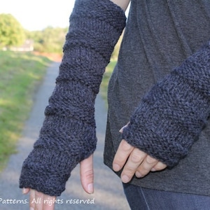 Könnte beinhalten: Ein Paar dunkelgraue gestrickte Armwärmer mit einem strukturierten Muster. Die Armwärmer werden von einer Person mit einem grauen Shirt und einer blauen Jeans getragen.