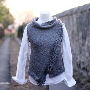 May include: A gray sleeveless sweater with a cowl neck and fringe detail. The sweater is displayed on a mannequin against a stone wall background.