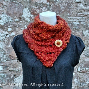 Pode incluir: Um cachecol de crochê em tons de laranja e vermelho com um botão de madeira. O cachecol é usado em um manequim contra um fundo de parede de pedra.
