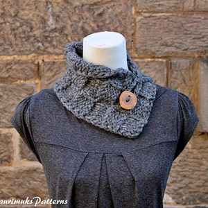 May include: A gray knitted cowl neck scarf with a wooden button. The scarf is worn on a gray shirt.