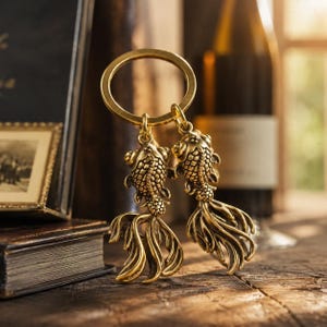 May include: A brass-toned metal keychain featuring two detailed goldfish charms suspended from a circular ring. Each fish charm displays intricate scale textures and long, flowing fins, crafted with a metallic finish that catches the light.