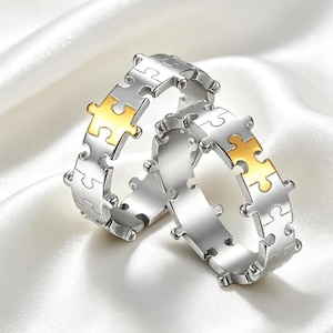 May include: Two silver-toned rings featuring a continuous puzzle piece pattern around the band. Each ring includes a single gold-toned puzzle piece accent integrated into the design. The rings are circular with interlocking edges.