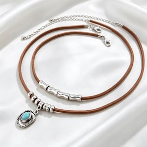 Puede incluir: Un conjunto de collares superpuestos con dos cordones de ante marrón. Un cordón incluye cuatro cuentas cilíndricas plateadas, mientras que el otro presenta un colgante ovalado central con una piedra turquesa y cuentas plateadas adicionales.