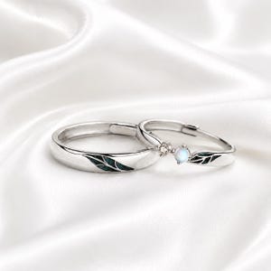 May include: Two silver-toned rings featuring a leaf-inspired design with dark green enamel inlay. One ring is a simple band, while the other is accented with a small, round, iridescent white stone and a tiny clear gemstone set in a silver prong.