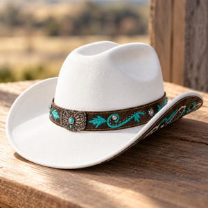 Può includere: Un cappello da cowboy in feltro bianco con una fascia in pelle marrone scuro decorata con ricami turchesi, borchie in metallo argentato e una fibbia centrale argentata con una piccola pietra turchese incastonata.