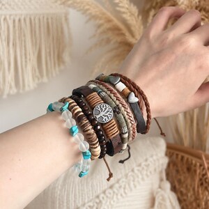 Puede incluir: Un conjunto de pulseras multicapa que incluye una hilera de cuentas blancas esmeriladas con acentos turquesa, cuentas de madera marrón oscuro, una banda de cuero marrón con un dije plateado de árbol de la vida, una banda tejida y un cordón trenzado.